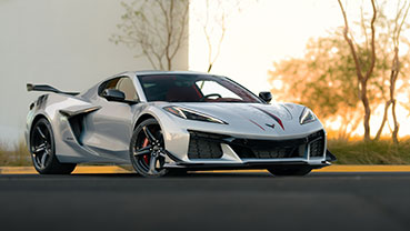 A silver sports car parked along a landscaped curb, shown from a front three‑quarter angle with plants and a light-colored building behind it.