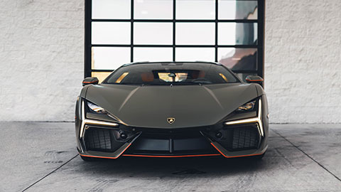 A dark gray supercar photographed head‑on in a modern garage space, showcasing sharp angular headlights and large front air vents.