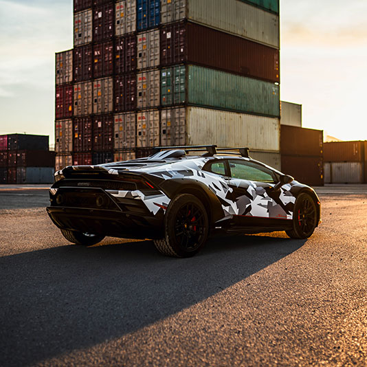 TESA white and black camouflaged sports car parked on pavement near stacked shipping containers at sunset.T
