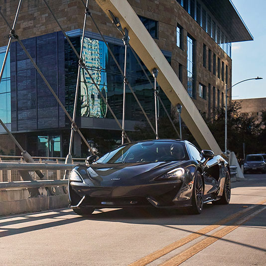 Black sports car driving across a modern suspension-style bridge in an urban area.