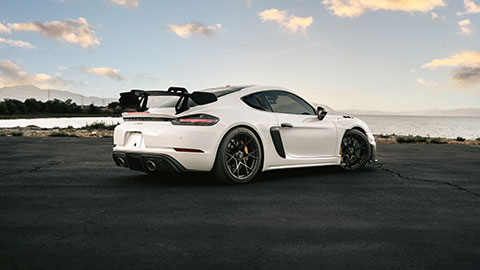 A white two‑door sports car parked on an empty paved area near a body of water, shown from the rear three‑quarter angle during sunset.