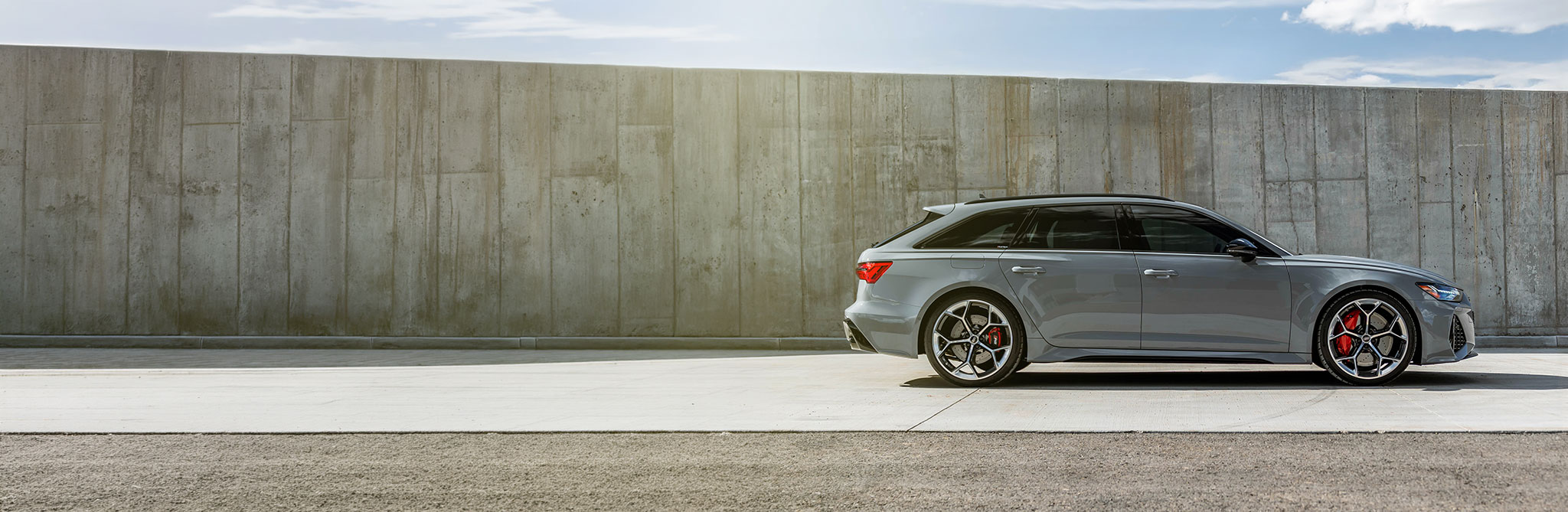 A gray performance wagon parked beside a tall concrete wall, shown from a side profile under bright daylight. 