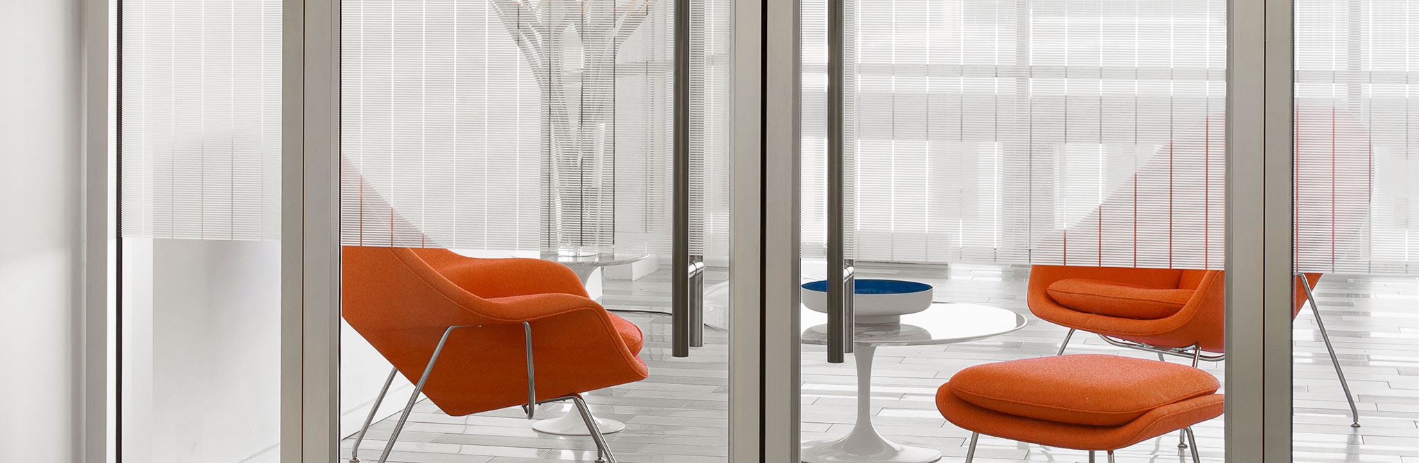 Modern office lounge with orange upholstered chairs and small white tables, viewed through glass walls with subtle horizontal privacy lines. 
